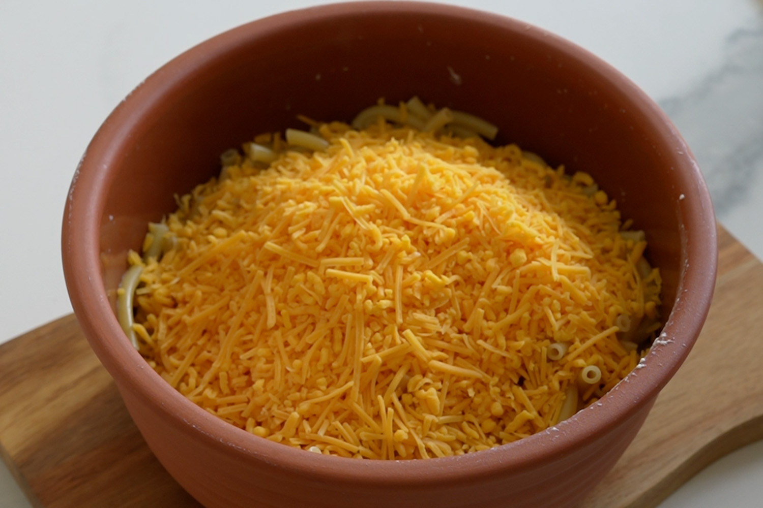 Schüssel mit geriebenem Cheddar-Käse auf Nudeln, auf einem Holzbrett stehend.