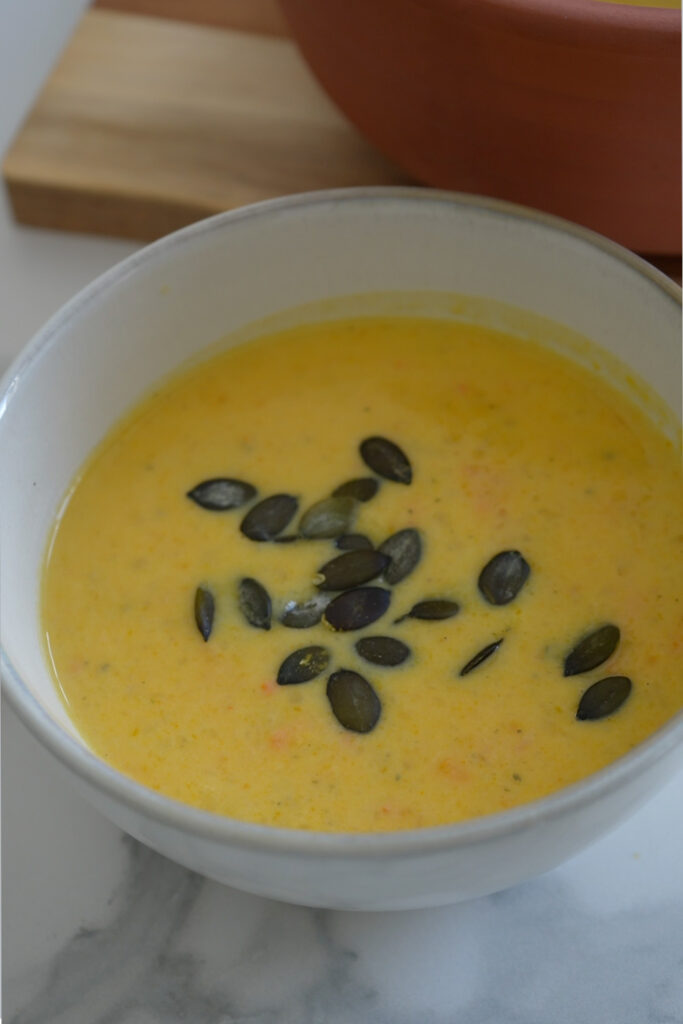 Weiße Schale mit gelber Kürbissuppe, garniert mit Kürbiskernen. Hintergrund aus Holz und Keramik.