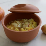 Terrakottatopf mit Gemüseeintopf, daneben zwei Kartoffeln und ein Kürbis auf weißem Hintergrund.