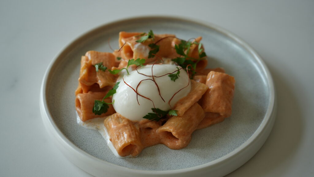 Rigatoni mit cremiger Sauce, garniert mit Petersilie und Safranfäden, serviert mit pochiertem Ei auf einem Teller.