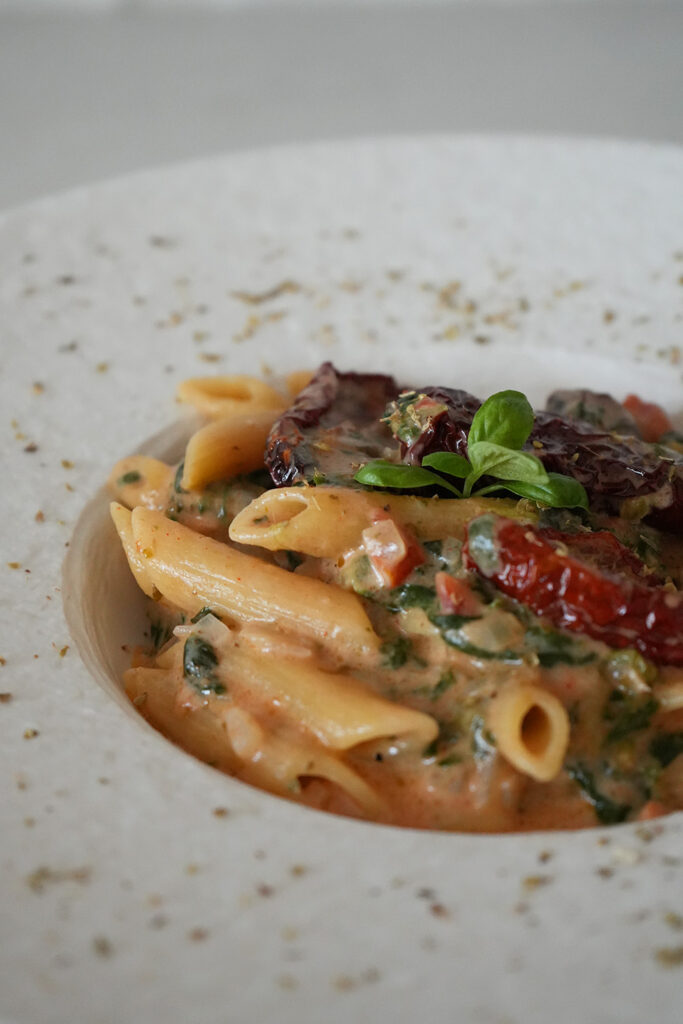Penne mit cremiger Tomaten-Sahne-Soße, getrockneten Tomaten und frischen Kräutern auf einem weißen Teller.