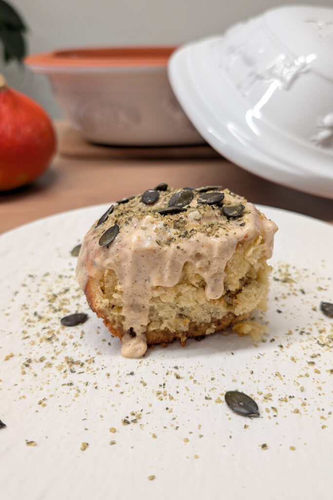 Ein Muffin mit glasierter Oberfläche und Kürbiskernen, umgeben von Krümeln auf einem Teller.