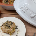 Ein Stück Gebäck mit Kürbiskernen auf einem Teller, daneben ein Keramiktopf und ein ganzer Kürbis.