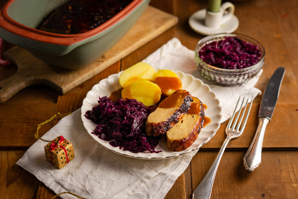 Teller mit veganem Braten, Kartoffeln, Rotkohl und Soße, daneben ein kleiner goldener Ziergegenstand.