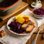 Teller mit veganem Braten, Kartoffeln, Rotkohl und Soße, daneben ein kleiner goldener Ziergegenstand.