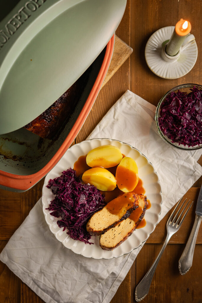 Teller mit Braten, Kartoffeln und Rotkohl. Daneben ein Topf und Kerze auf Holz.