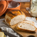 Hausgemachtes Brot auf Holzbrett, daneben Butter und ein Messer, im Hintergrund ein Topf und ein Glas mit Sauerteig.