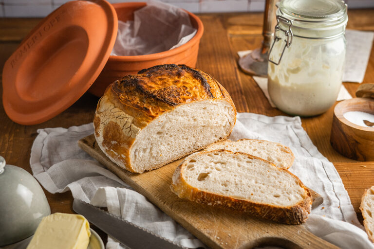 Hausgemachtes Brot auf Holzbrett, daneben Butter und ein Messer, im Hintergrund ein Topf und ein Glas mit Sauerteig.
