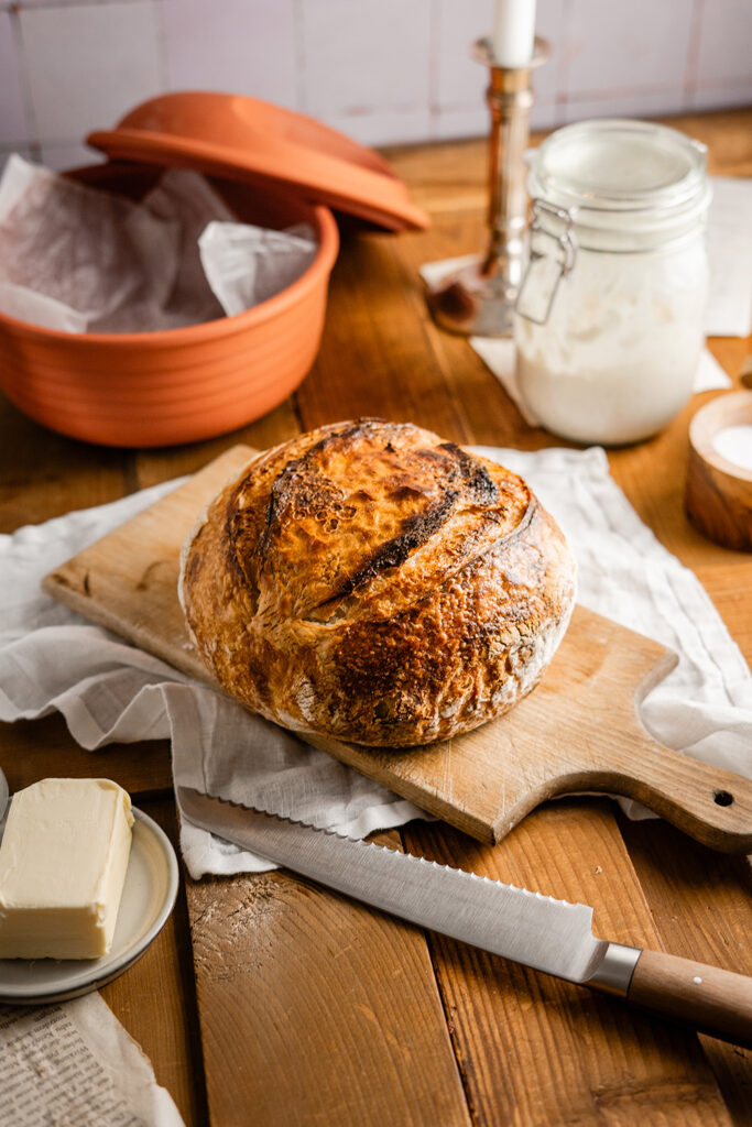 Hausgemachtes Brot auf Holzbrett, daneben Butter und ein Messer, im Hintergrund ein Topf und ein Glas mit Sauerteig.