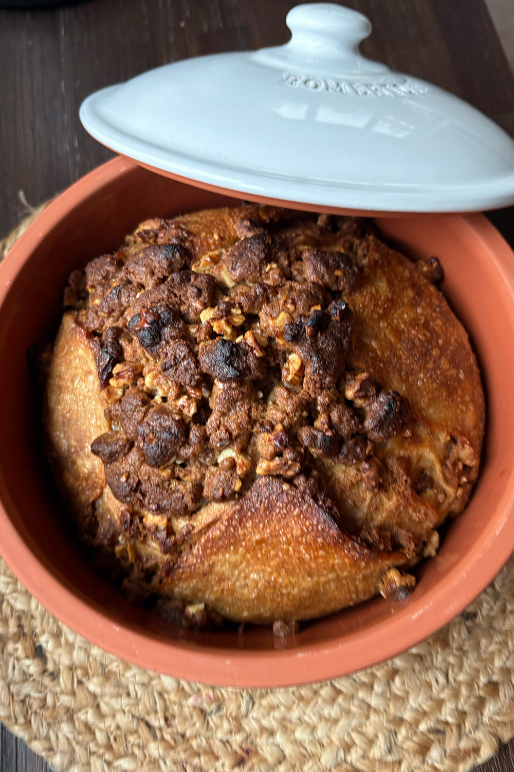 Brauner Laib Brot in einem runden, braunen Topf mit leicht geöffnetem weißen Deckel.