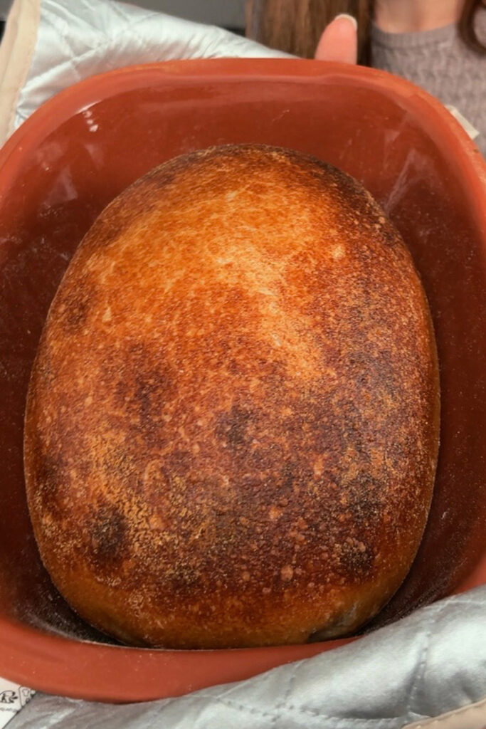 Frisch gebackenes, goldbraunes Brot in einem roten Keramiktopf auf einem gepolsterten Ofenhandschuh.