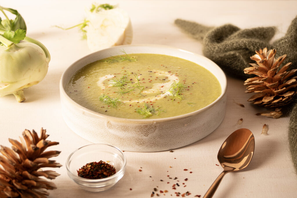 Schale mit grüner Suppe, garniert mit Kräutern und Gewürzen, daneben Kohlrabi, Gewürzglas, Tannenzapfen und Löffel.