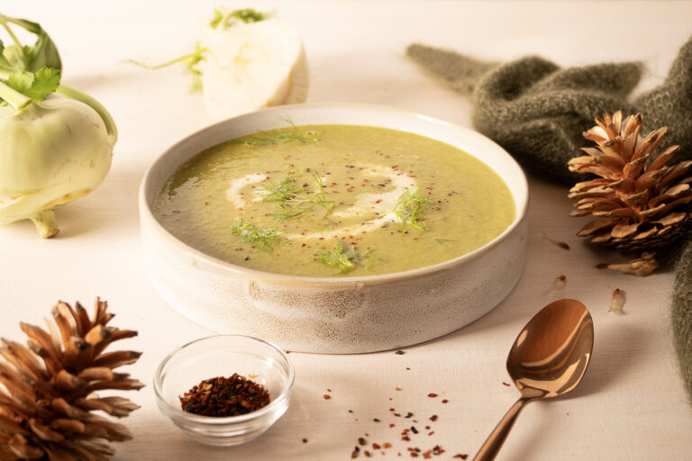 Schale mit grüner Suppe, garniert mit Kräutern und Gewürzen, daneben Kohlrabi, Gewürzglas, Tannenzapfen und Löffel.