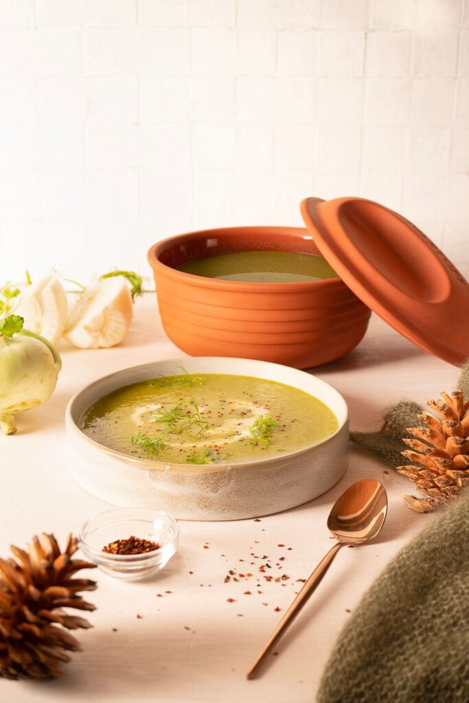 Schüssel mit cremiger, grüner Suppe, garniert mit Kräutern, umgeben von Gewürzen und einem Löffel auf hellem Hintergrund.