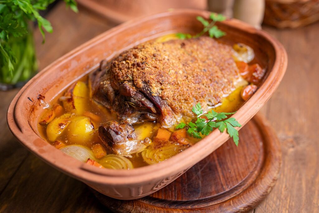 Gebratenes Fleisch mit Kruste in einem Tontopf, umgeben von Kartoffeln, Karotten und Zwiebeln, dekoriert mit Petersilie.