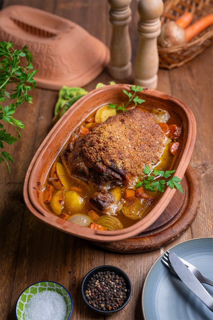 Braten mit Kruste in einem Römertopf, umgeben von Gemüse, auf einem Holztisch mit Gewürzen und Besteck.