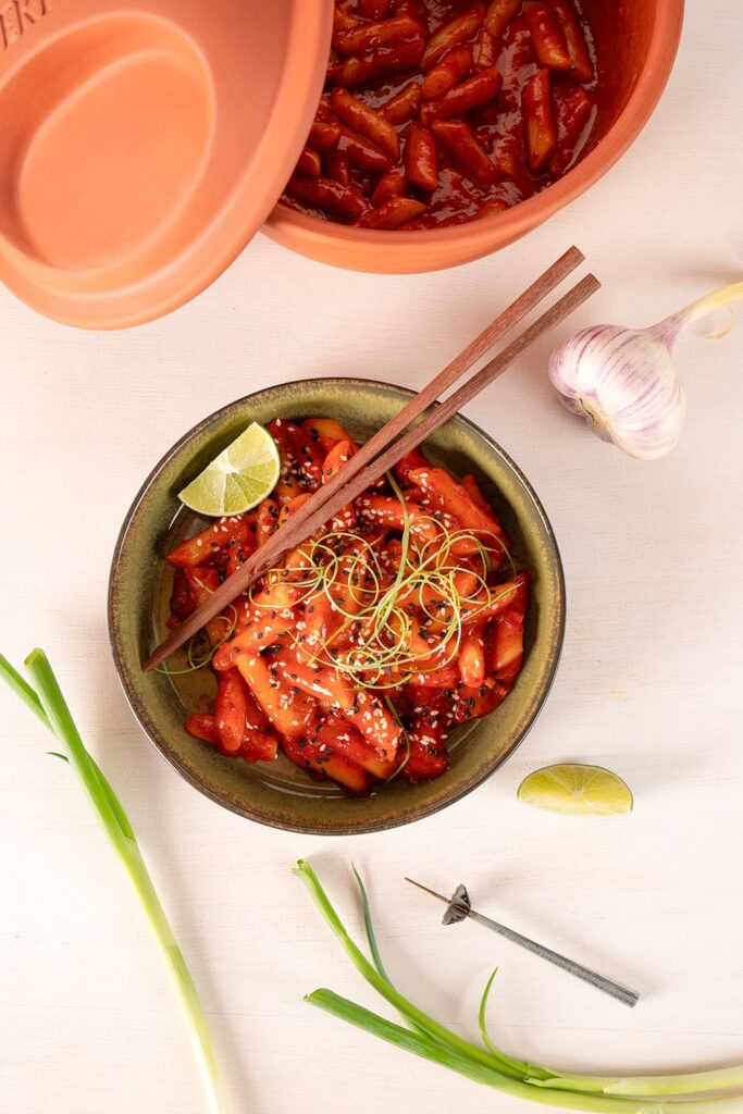 Koreanisches Tteokbokki in Schale mit Stäbchen, Limette, Knoblauch und Frühlingszwiebeln auf hellem Hintergrund.
