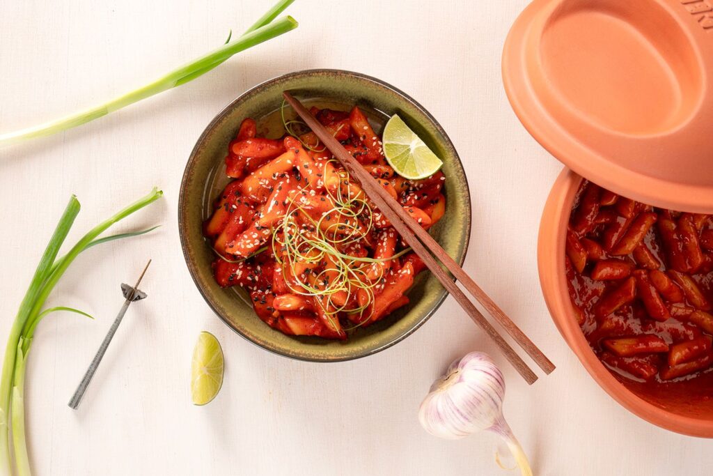 Schale mit Tteokbokki, garniert mit Frühlingszwiebeln und Sesam, daneben Knoblauch, Limette und Essstäbchen auf weißem Tisch.