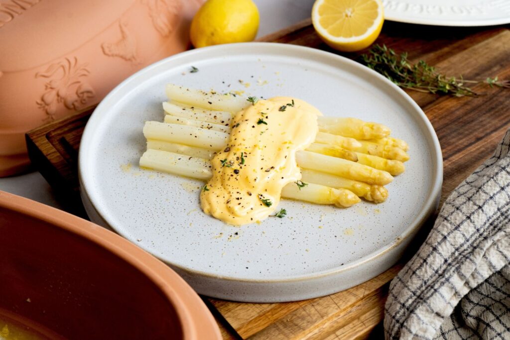 Ein Teller mit weißem Spargel, bedeckt mit einer gelben Soße, dekoriert mit Kräutern und einer halbierten Zitrone im Hintergrund.