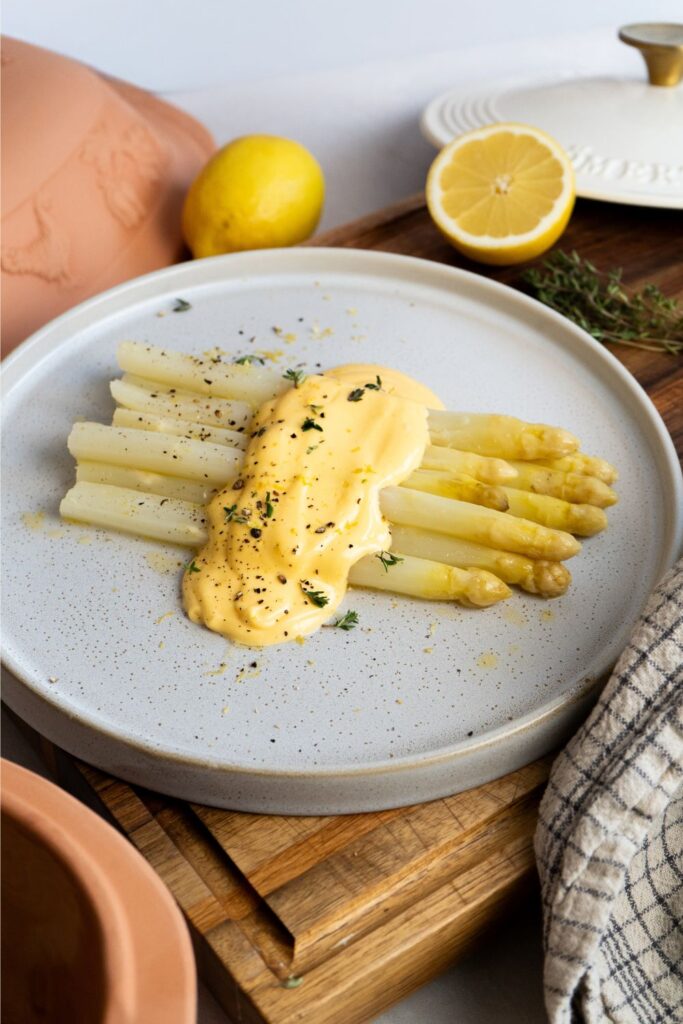Weißer Spargel mit Sauce Hollandaise, auf einem Teller serviert, garniert mit Pfeffer und Kräutern. Halbierte Zitrone im Hintergrund.
