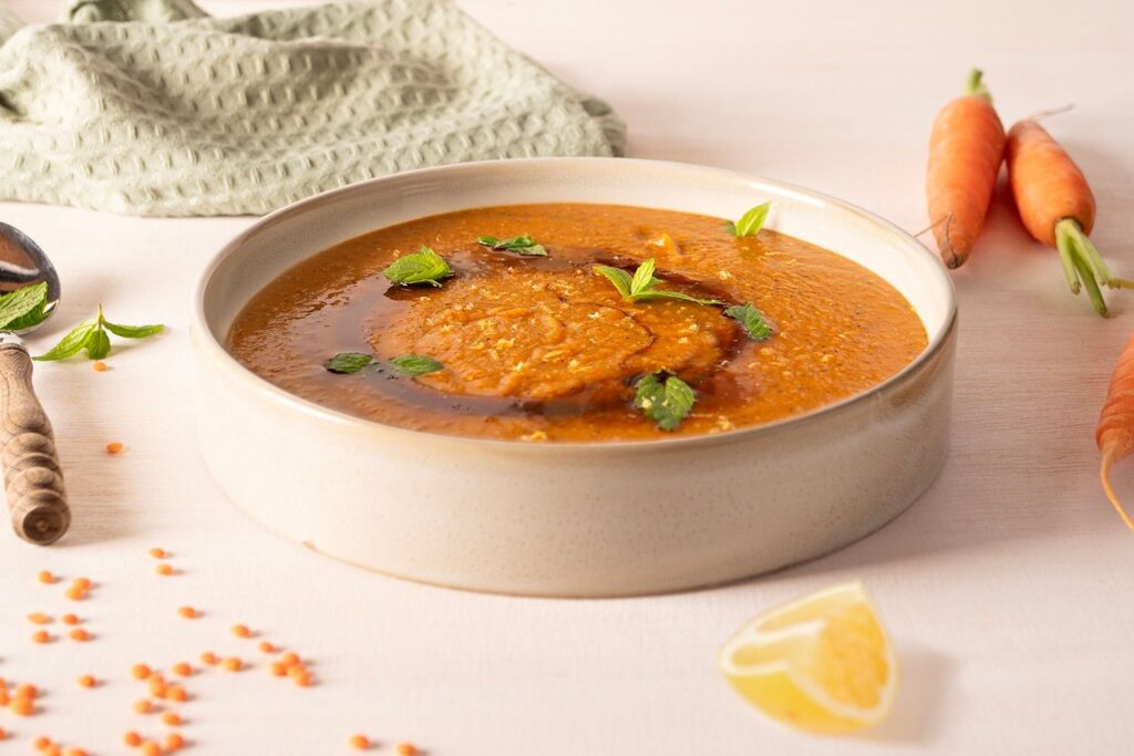 Schüssel mit Karotten-Linsensuppe, garniert mit Minzblättern. Im Hintergrund frische Karotten und Zitronenspalten.