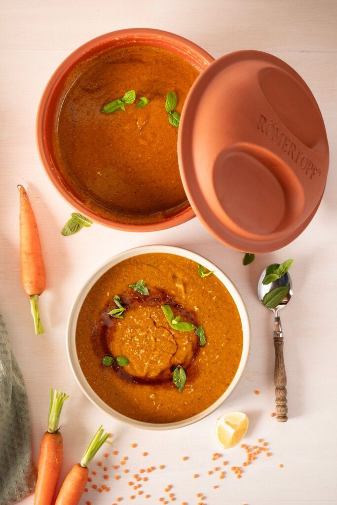 Suppe in brauner Schale mit Minzblättern, daneben Karotten, Linsen und ein Löffel auf hellem Hintergrund.