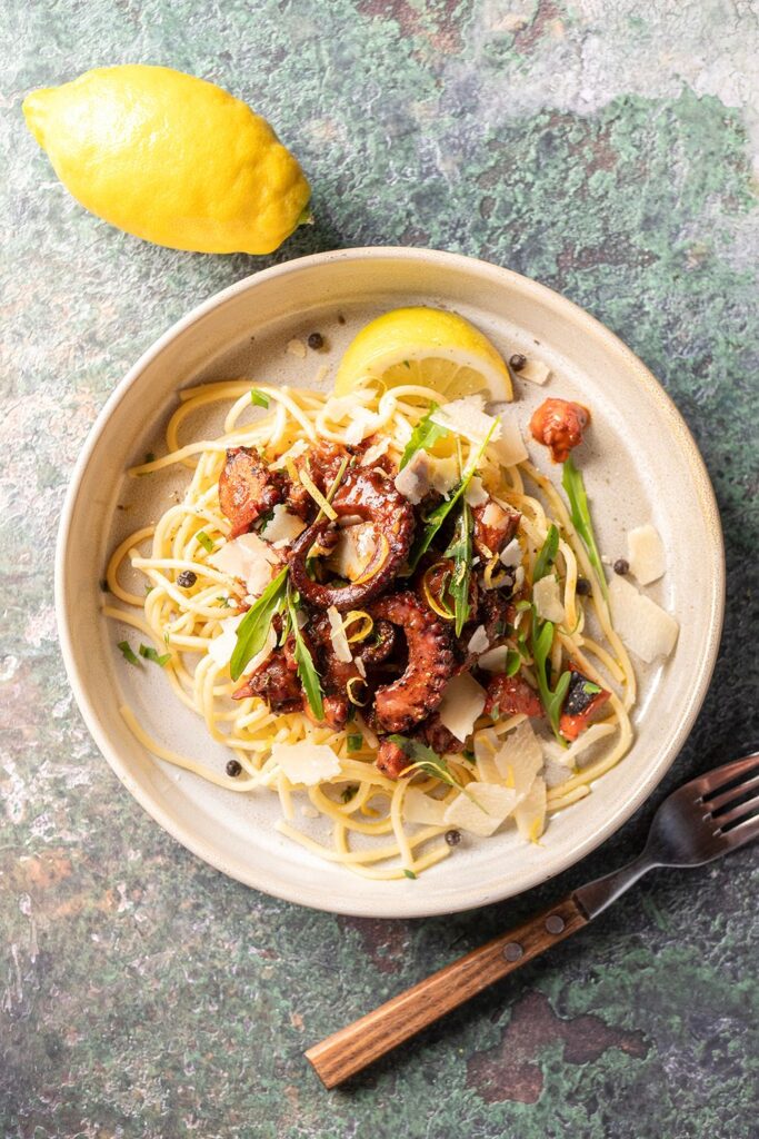 Teller mit Spaghetti, Oktopus, Parmesan und Rucola. Daneben eine Zitrone und eine Gabel auf einer grünen Oberfläche.