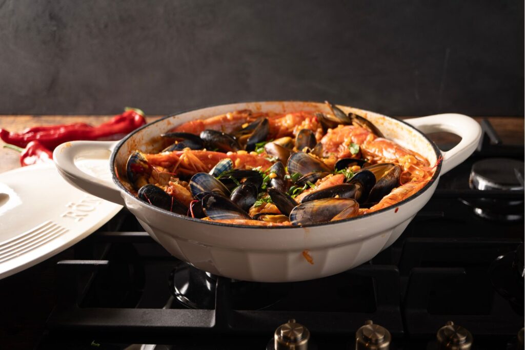 Weißer Topf mit Meeresfrüchte-Paella, Muscheln und Garnelen auf dem Herd, mit roten Paprikaschoten im Hintergrund.