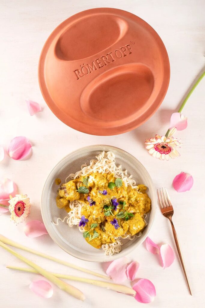 Terrakotta-Römertopf neben Teller mit Garnelen-Curry auf Nudeln, dekoriert mit rosa Blüten und Zitronengras.