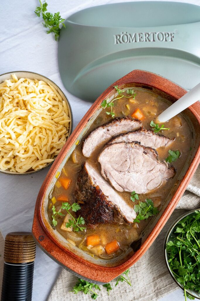 Braten im Römertopf mit Soße, Karotten und Petersilie, daneben eine Schale mit Spätzle und frische Petersilie.
