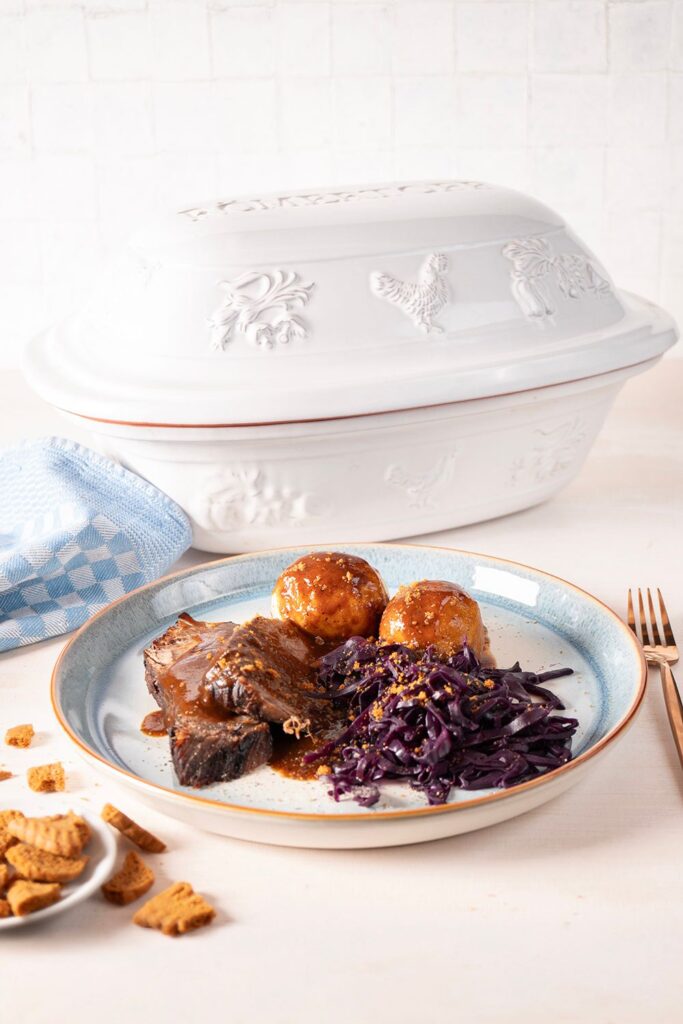 Teller mit Braten, Rotkohl und Knödeln, serviert neben einem weißen Römertopf und blauem Tuch auf hellem Tisch.