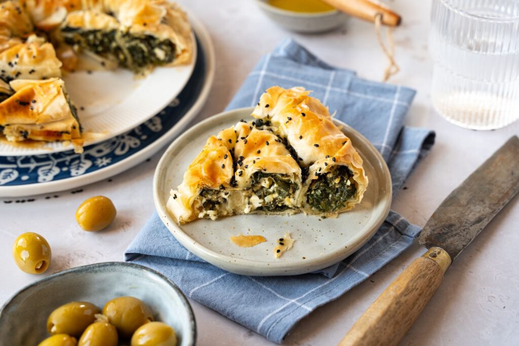 Spinat-Feta-Börek auf einem Teller, mit schwarzem Sesam bestreut. Daneben Oliven und ein Messer auf einem blauen Tuch.