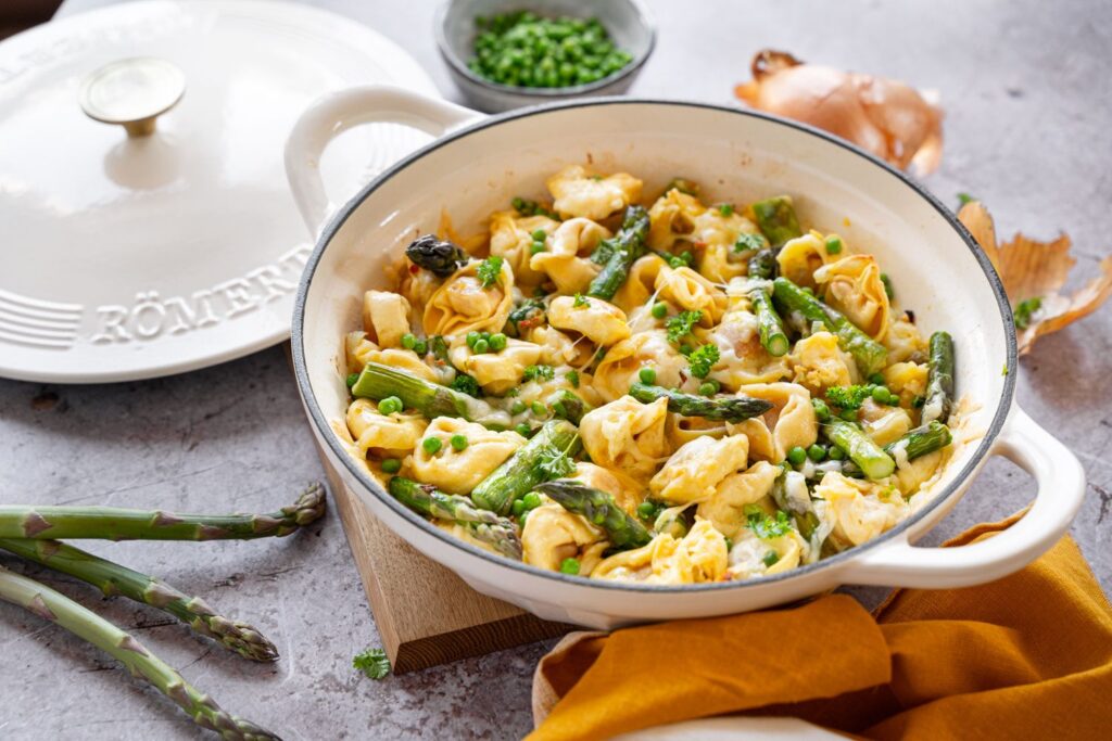 Weiße Auflaufform mit Tortellini, grünem Spargel, Erbsen und Kräutern auf einem Tisch, daneben ein weißer Topfdeckel.