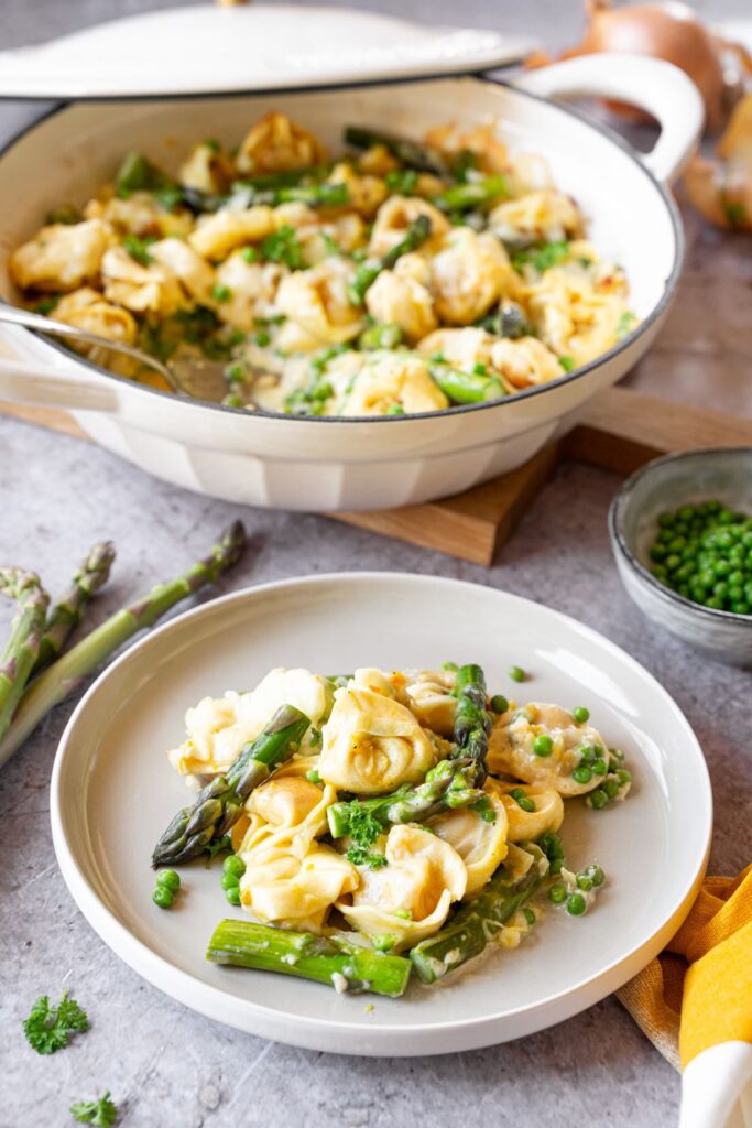 Tortellini mit grünem Spargel und Erbsen in cremiger Sauce, serviert auf einem weißen Teller und im weißen Topf.