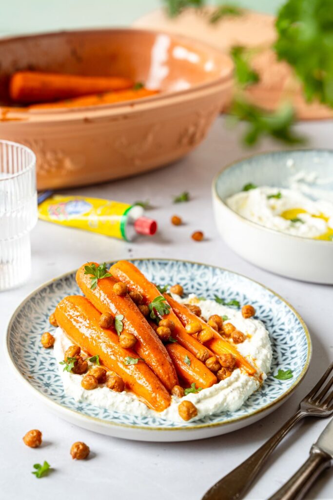 Glasierte Honig-Harissa-Möhren auf Whipped Feta mit Kichererbsen und Petersilie, im Hintergrund der Römertopf Rustico