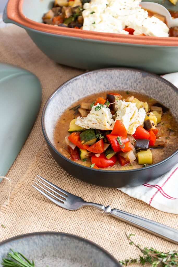 Portion Ratatouille mit cremigem Feta in einer dunklen Schüssel, im Hintergrund der grüne Römertopf Culinario
