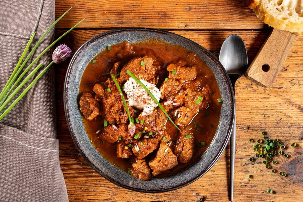 Schwarzbier-Gulasch in einer dunklen Schüssel auf Holztisch, mit Schmand, Schnittlauch und Chiliflocken getoppt, daneben frisches Brot