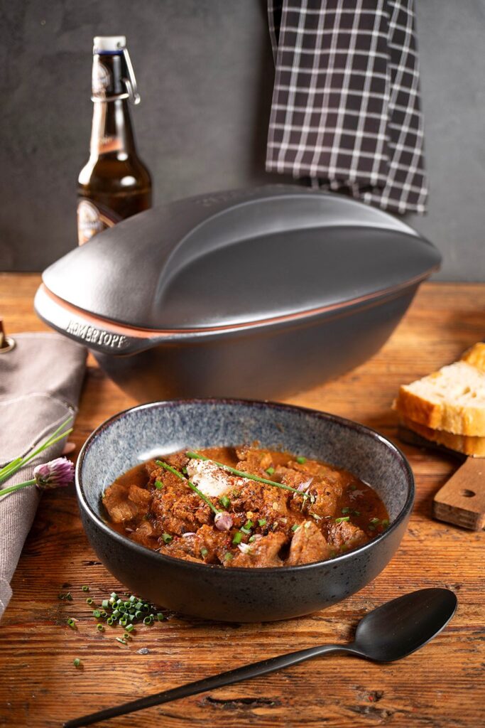 Portioniertes Schwarzbier-Gulasch in dunkler Schale, im Hintergrund der schwarze Römertopf und eine Schwarzbierflasche