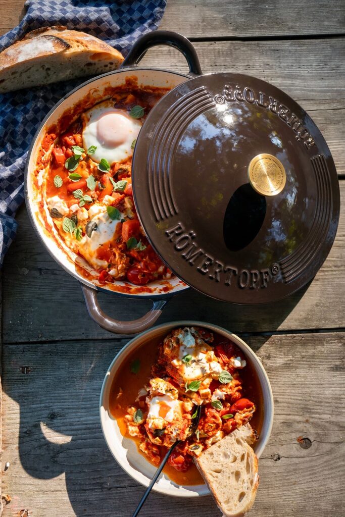 Shakshuka mit Feta und Eiern in der dunklen Römertopf Schmorpfanne von oben, daneben eine Schüssel mit der fertigen Portion