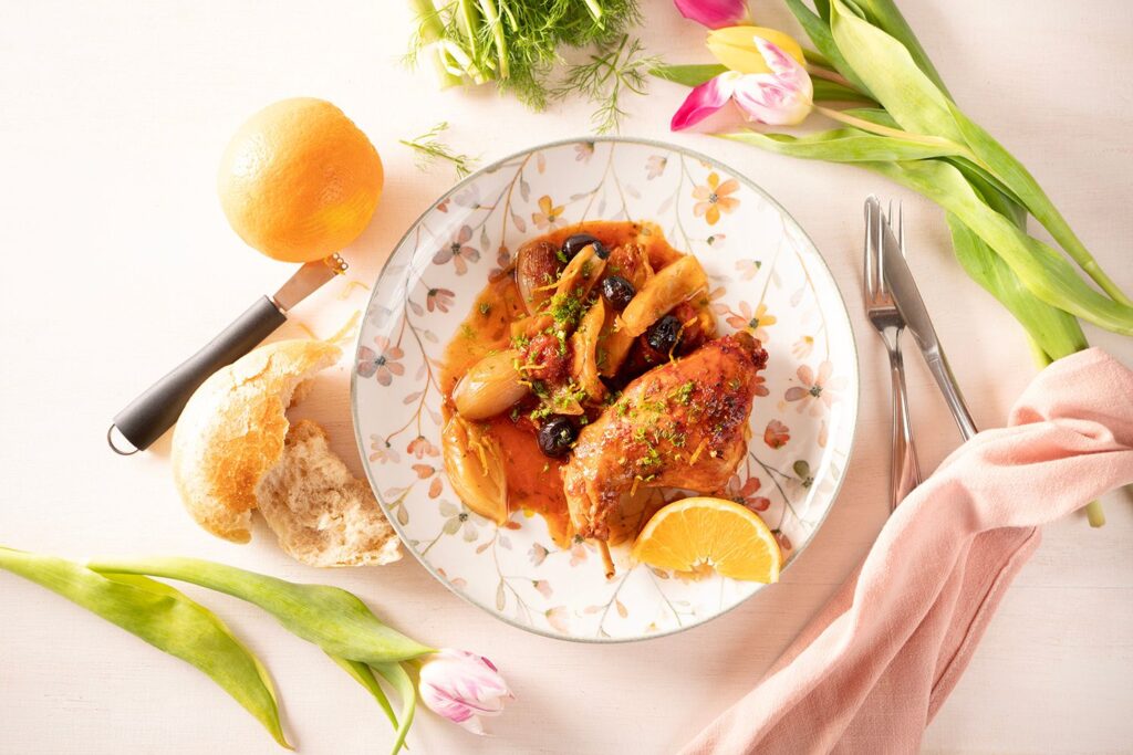 Kaninchen mit Orangen, Fenchel und schwarzen Oliven in aromatischer Tomatensauce auf einem geblümten Teller von Moritz und Moritz, dazu Weißbrot und Orangenscheibe