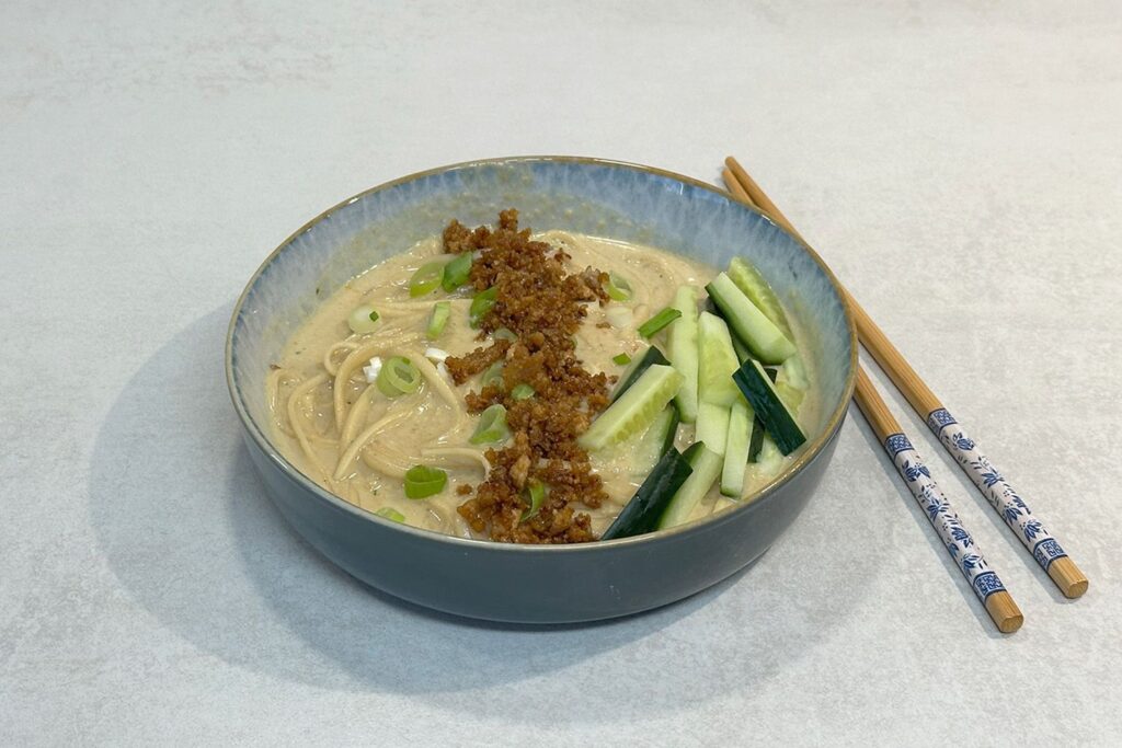 Ramen mit Crispy Tofu in cremiger Kokos-Erdnuss-Brühe, getoppt mit Frühlingszwiebeln und Gurkenstreifen in einer blauen Schüssel
