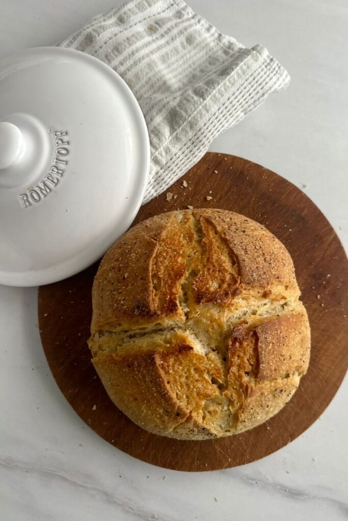 Dinkel Sauerteigbrot mit Pfefferkruste von oben auf Holzbrett, daneben weißer Römertopf Brottopf-Deckel