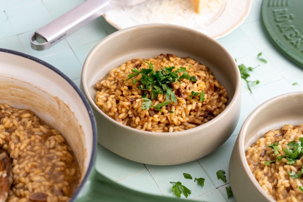Cremiges Steinpilzrisotto von oben in zwei Schüsseln und der grünen Stielkasserolle, mit Parmesan, Reibe und Pfeffermühle