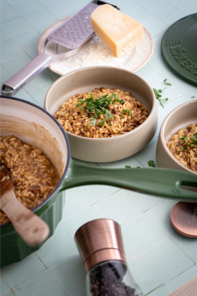Steinpilzrisotto in einer cremefarbenen Schüssel mit frischer Petersilie, daneben die grüne Römertopf Stielkasserolle und Parmesan
