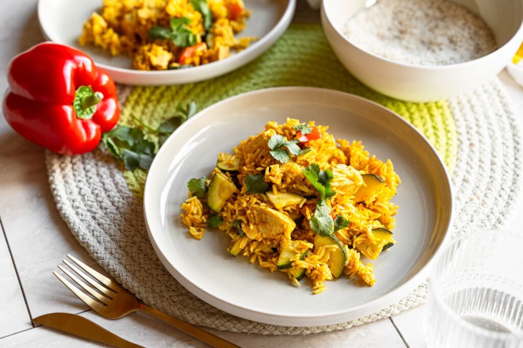 Zucchini Curry mit Hähnchen und Reis auf weißem Teller, garniert mit frischem Koriander, im Hintergrund rote Paprika
