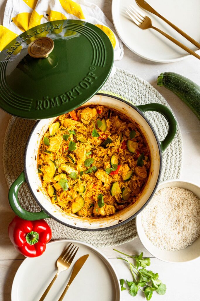 Grüner Römertopf Gusseisentopf mit geöffnetem Deckel und angerichtetem Zucchini Curry, daneben rohe Zucchini und Reis