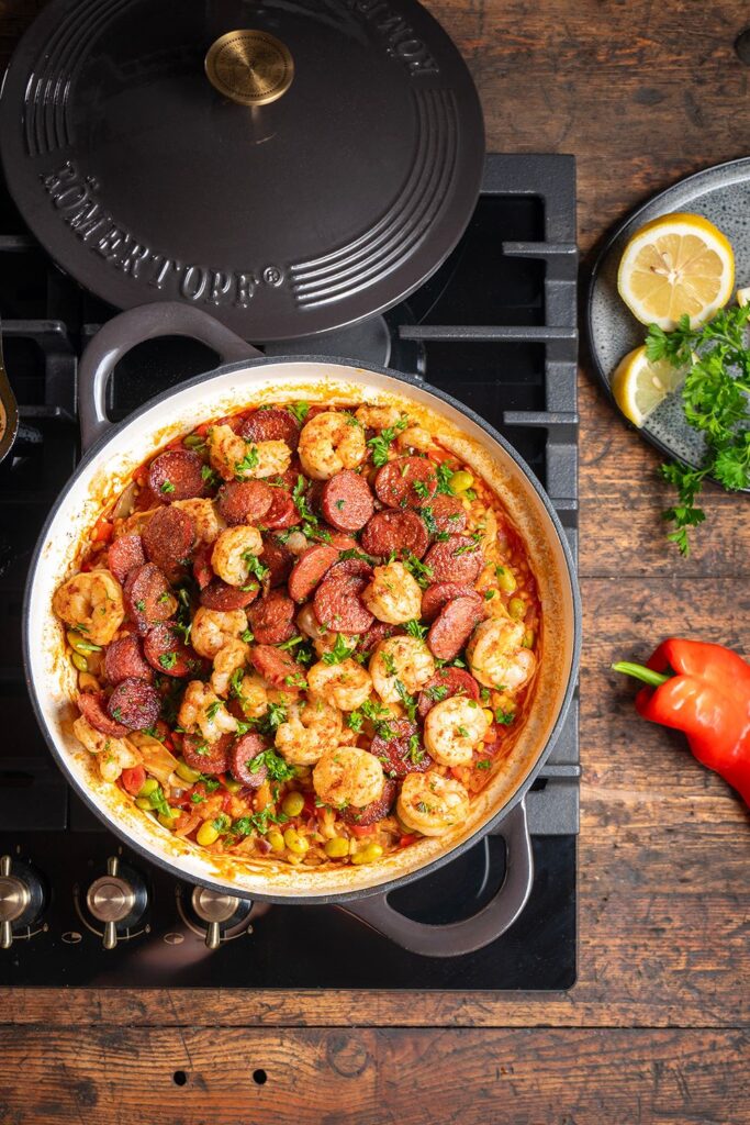 Bild 1: Paella mit Garnelen und Sucuk in schwarzer Schmorpfanne auf dem Herd, garniert mit Petersilie, daneben Zitronenscheiben und rote Paprika