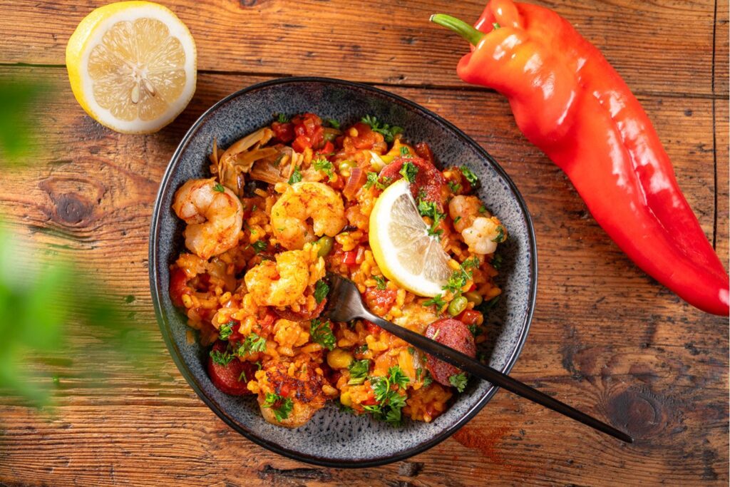 Bild 2: Angerichtete Paella mit Garnelen, Sucuk und Zitronenspalte in Moritz und Moritz Schale, im Hintergrund rote Paprika und halbe Zitrone