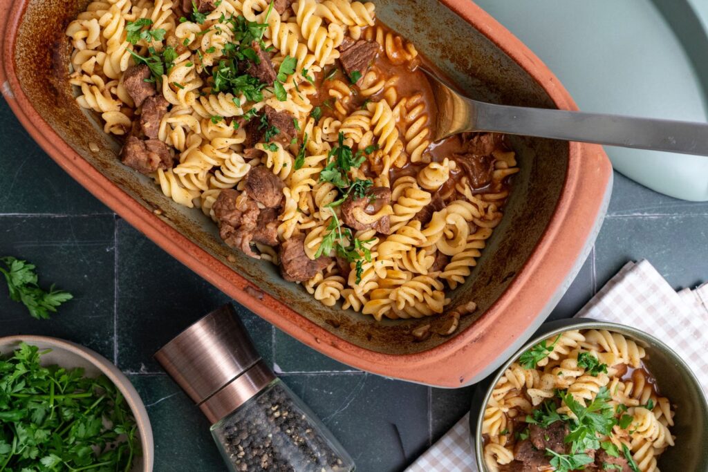 Bild 1: Gulasch-Pasta-Topf mit Rindfleisch und Muschelnudeln im Römertopf Culinario, garniert mit Petersilie, im Hintergrund eine Portion in grüner Schale
