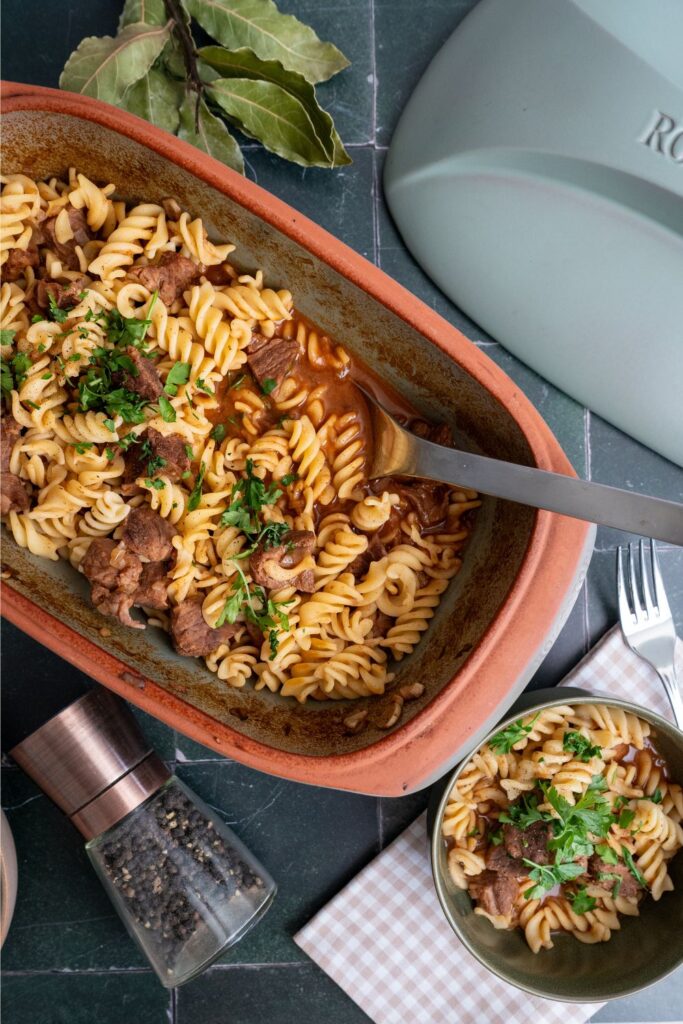 Bild 2: Gulasch-Pasta-Topf mit Rindfleisch und Muschelnudeln in Terrakotta-Bräter, umgeben von Lorbeerblättern und Pfeffermühle